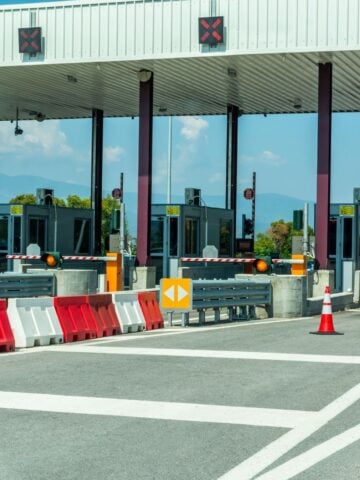 A closed toll booth with red X signs above the lanes, barriers blocking entry, and traffic cones placed in front. The sky is clear and there are trees in the background.