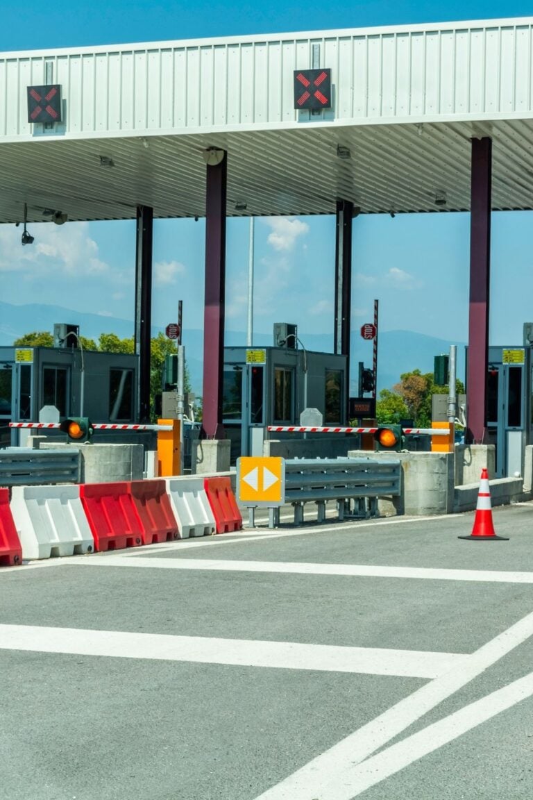 A closed toll booth with red X signs above the lanes, barriers blocking entry, and traffic cones placed in front. The sky is clear and there are trees in the background.