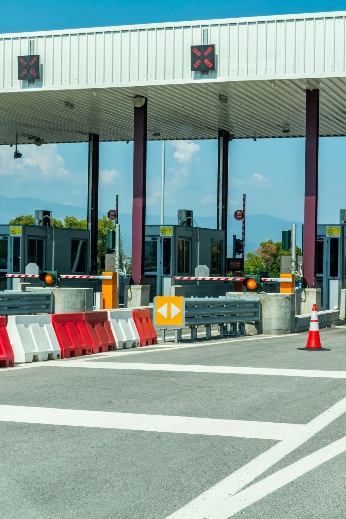A closed toll booth with red X signs above the lanes, barriers blocking entry, and traffic cones placed in front. The sky is clear and there are trees in the background.