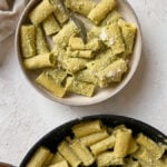 A bowl and pan of rigatoni pasta coated in green pesto sauce, with a fork in the bowl and a serving spoon in the pan, on a light textured surface.