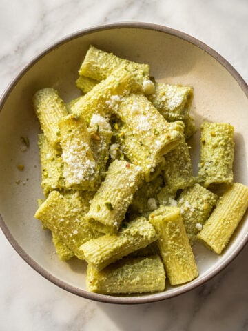 A bowl of rigatoni pasta coated in green pesto sauce, topped with a light sprinkle of grated cheese, sits on a white marble surface.