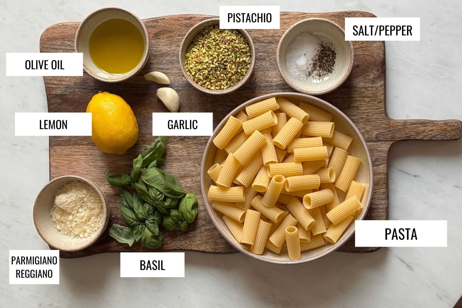 A wooden board displays labeled ingredients: a bowl of rigatoni pasta, chopped pistachios, salt and pepper, olive oil, lemon, garlic, grated Parmigiano Reggiano, and fresh basil leaves.