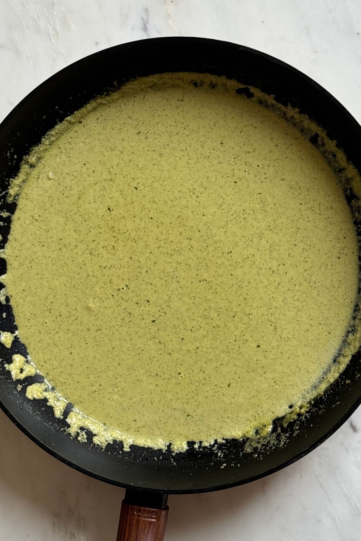 A black pan with creamy green sauce spread evenly inside, sitting on a white marble surface.