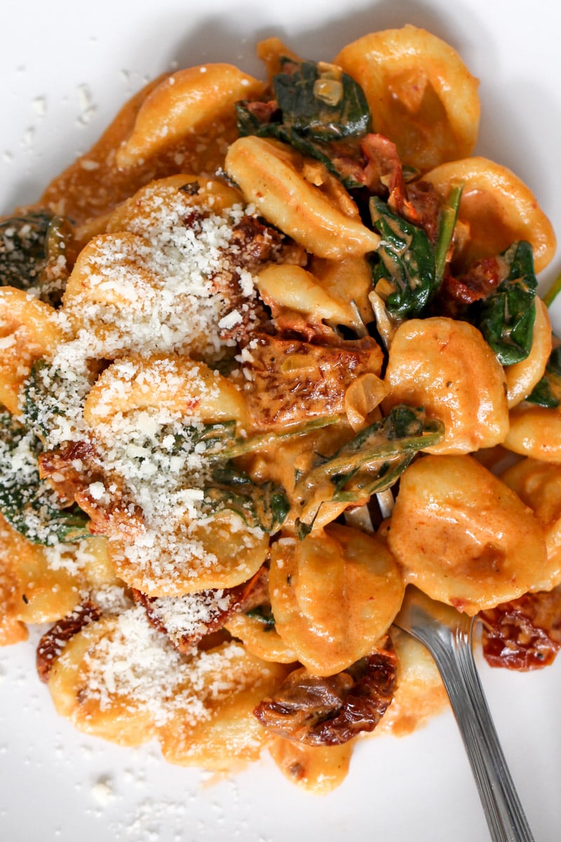 Creamy orecchiette pasta with spinach, sun-dried tomatoes, and grated parmesan cheese on top, served on a white plate with a fork.