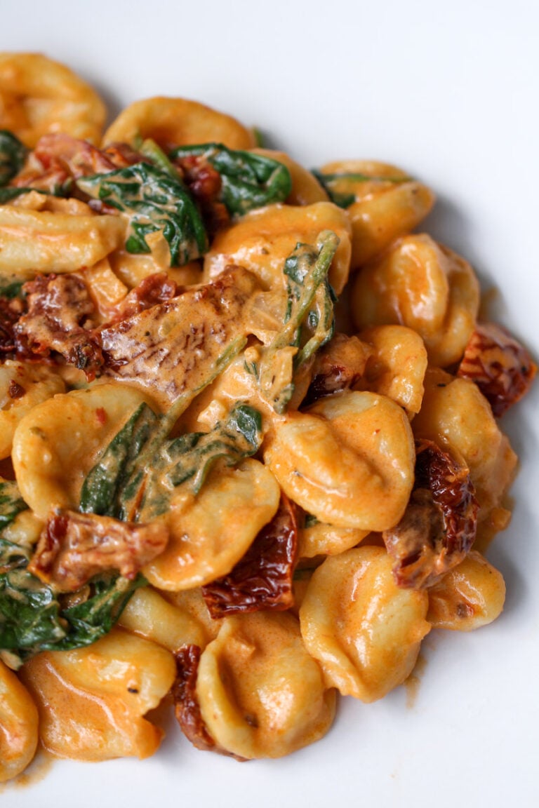 Close-up of creamy pasta with orecchiette, wilted spinach, sun-dried tomatoes, and pieces of seared chicken, all coated in a rich orange sauce on a white plate.