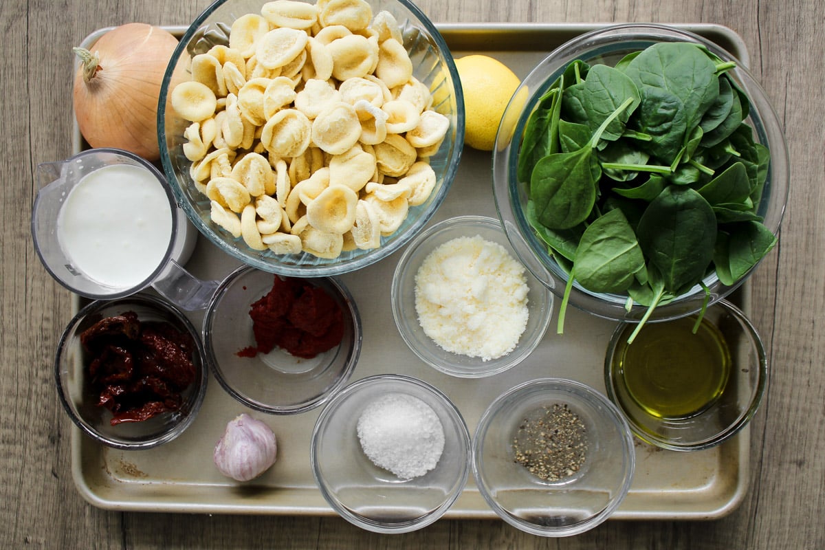 Assorted ingredients in bowls and cups on a tray, including orecchiette pasta, fresh spinach, lemon, onion, cream, sun-dried tomatoes, tomato paste, garlic, Parmesan, olive oil, salt, and pepper.