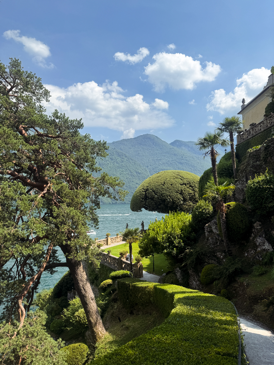 A scenic garden with manicured bushes, trees, and a path overlooking a blue lake, with mountains and a partly cloudy sky in the background.