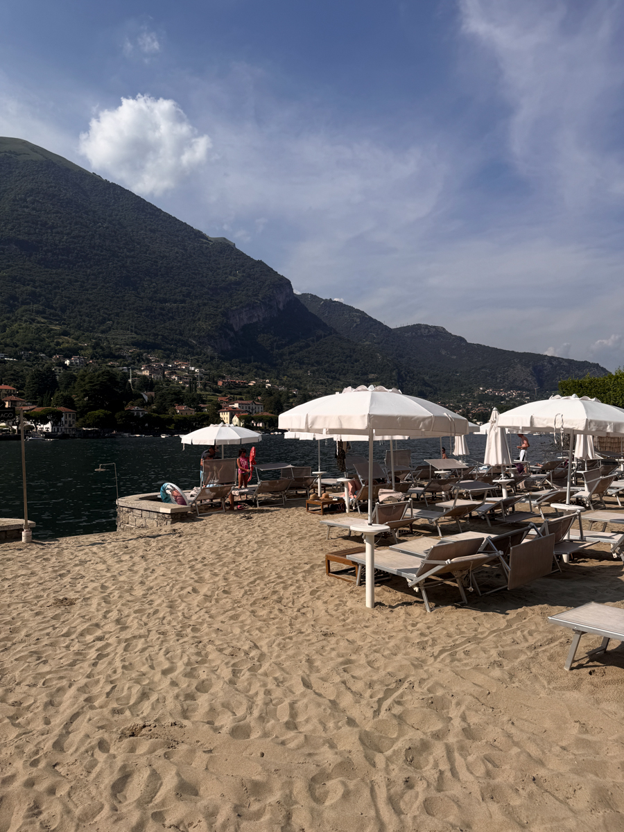 Sandy beach with white umbrellas and lounge chairs by a lake, mountains in the background, and people relaxing near the water under a partly cloudy sky.