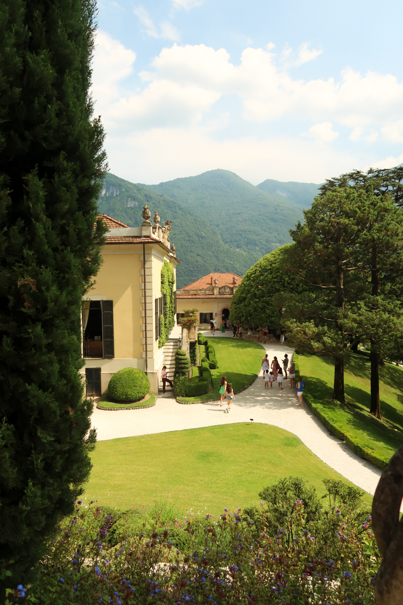 A yellow villa with green shutters sits amid manicured lawns, tall trees, and mountains in the background. People walk along a curved gravel path under a partly cloudy sky.