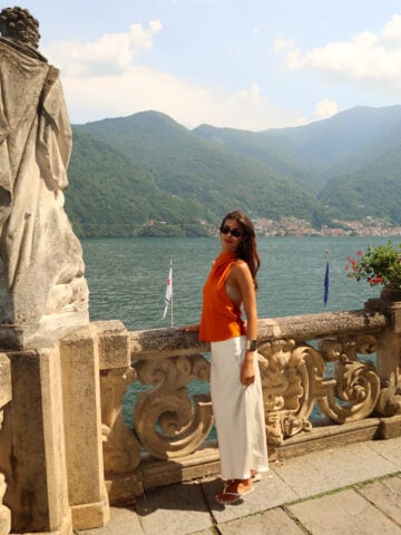 A woman in an orange top and white skirt stands by a stone railing overlooking a lake with mountains in the background on a sunny day.