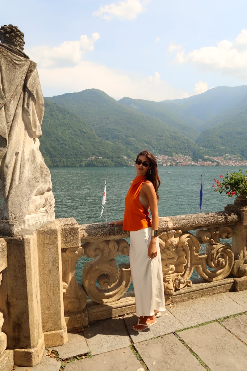 A woman in an orange top and white skirt stands by a stone railing overlooking a lake with mountains in the background on a sunny day.