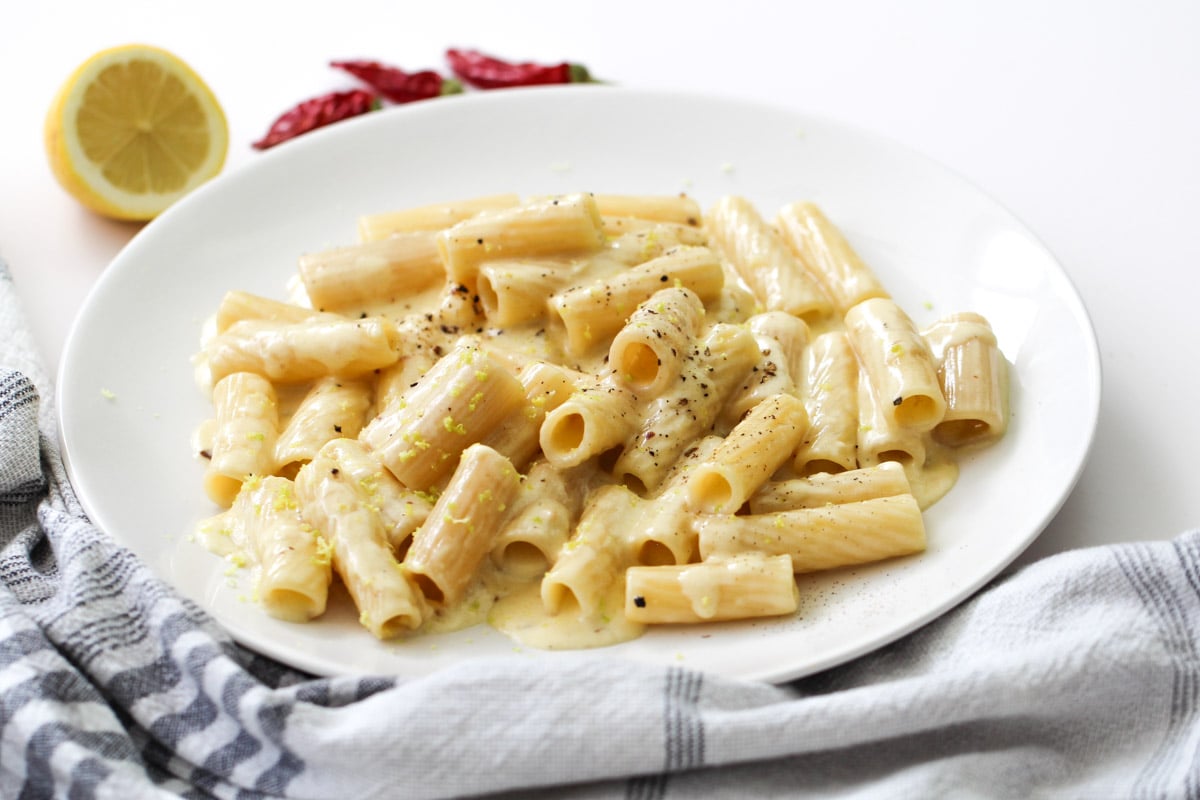 A plate of rigatoni pasta in a creamy white sauce, garnished with black pepper and lemon zest, sits on a white table with a lemon half, red chili peppers, and a striped cloth nearby.