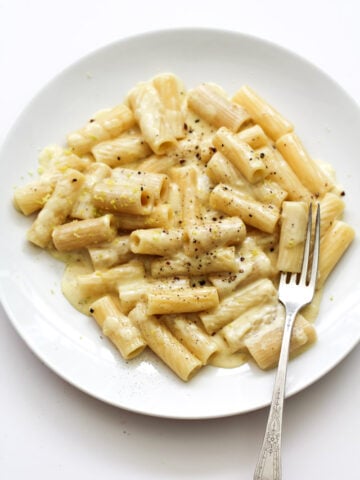 A white plate with rigatoni pasta in a creamy white sauce, topped with cracked black pepper, with a fork resting on the side of the plate.