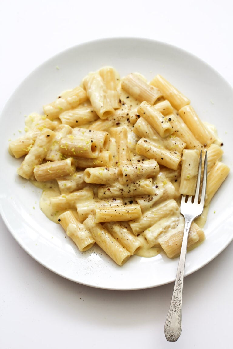 A white plate with rigatoni pasta in a creamy white sauce, topped with cracked black pepper, with a fork resting on the side of the plate.