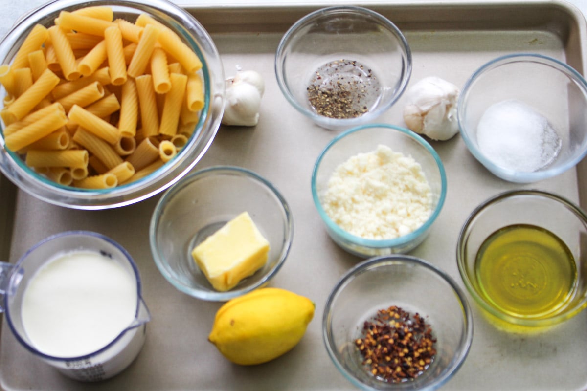 A tray holds bowls of rigatoni pasta, grated cheese, milk, butter, lemon, olive oil, red pepper flakes, salt, pepper, and two garlic bulbs.