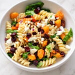A white bowl of rotini pasta salad with roasted squash, dried cranberries, spinach, pine nuts, and crumbled feta cheese.