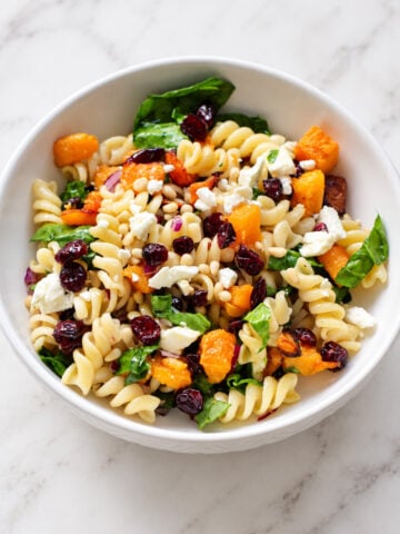 A white bowl of rotini pasta salad with roasted squash, dried cranberries, spinach, pine nuts, and crumbled feta cheese.