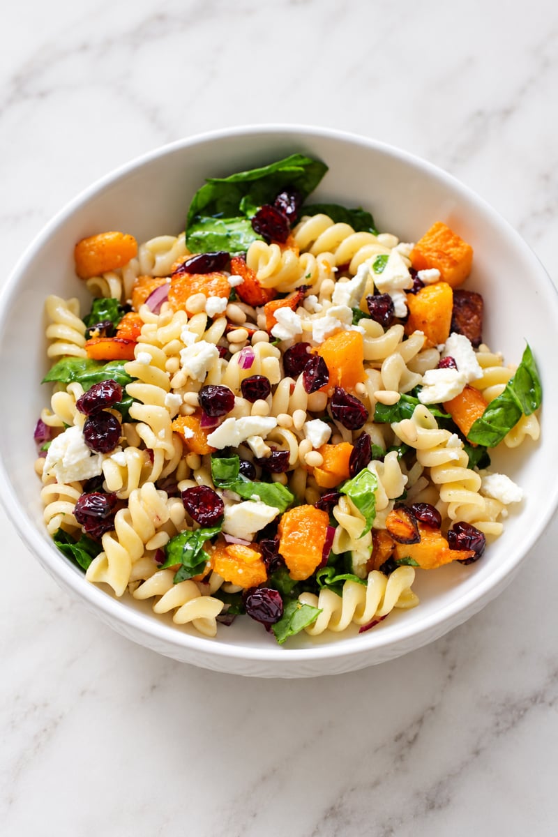 A white bowl of rotini pasta salad with roasted squash, dried cranberries, spinach, pine nuts, and crumbled feta cheese.