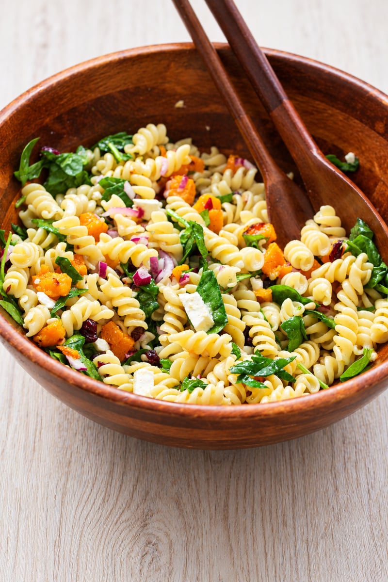 A wooden bowl of rotini pasta salad with roasted squash, spinach, red onion, and crumbled cheese, served with wooden salad spoons.