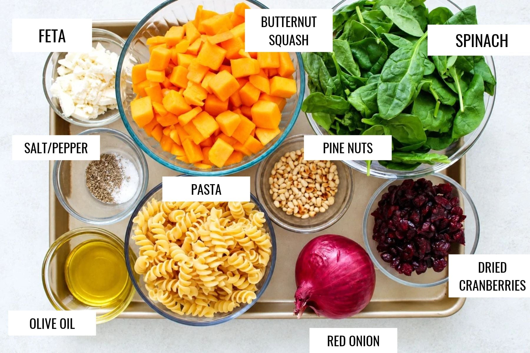 Overhead view of a tray holding bowls of ingredients: feta, diced butternut squash, spinach, salt and pepper, pine nuts, pasta, olive oil, a red onion, and dried cranberries.