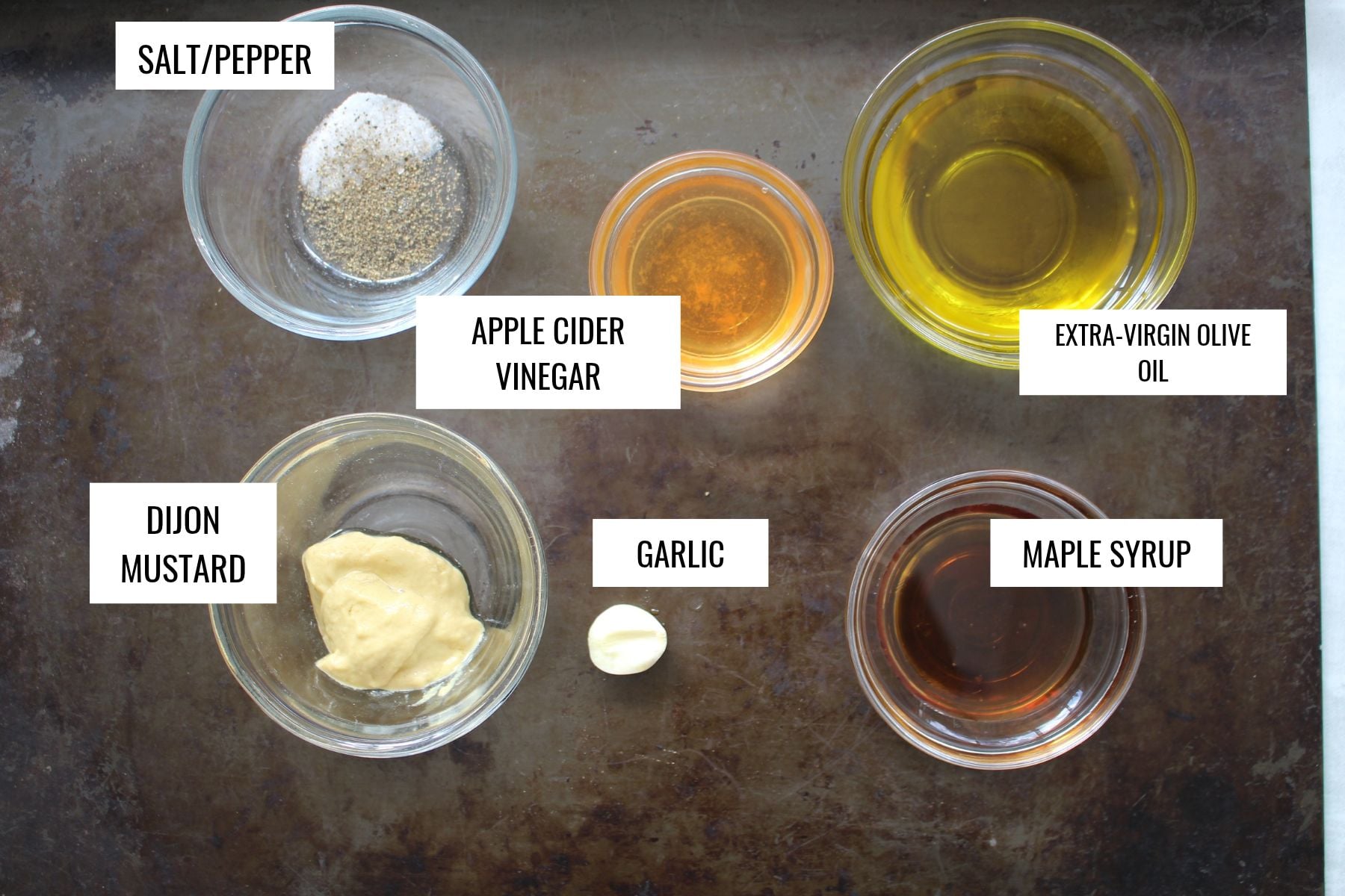 Six glass bowls containing salt and pepper, apple cider vinegar, extra-virgin olive oil, Dijon mustard, maple syrup, and a single clove of garlic, arranged on a dark metal tray with identifying labels for each ingredient.