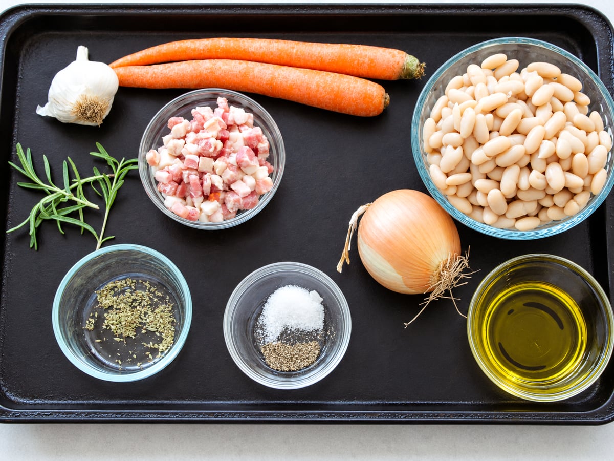 Ingredients for a meal on a baking sheet: two carrots, a head of garlic, diced bacon, rosemary, dried herbs, salt, pepper, a white onion, white beans in a glass bowl, and olive oil.