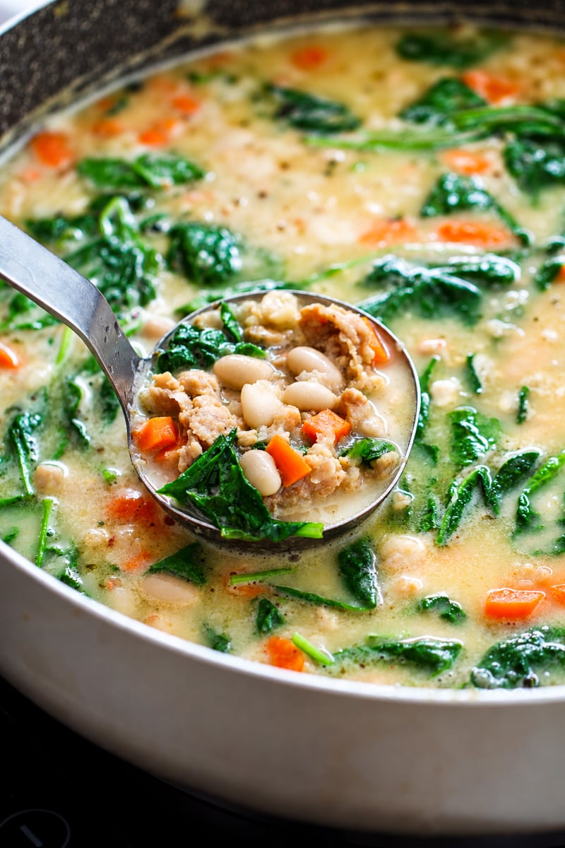 A ladle holds a scoop of creamy soup containing white beans, sausage, and carrots above a pan of the same soup with spinach.