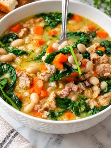 A spoon lifts a portion of creamy soup containing white beans, sausage, carrots, and spinach from a full bowl.