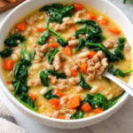 A bowl of creamy soup with ground meat, white beans, carrots, and spinach, with a spoon resting in the bowl.